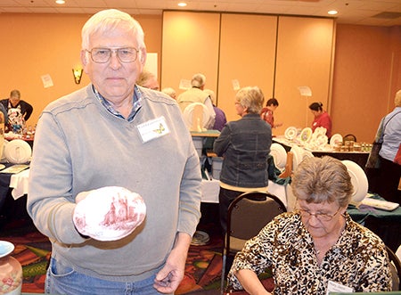 Carl Grandstrand holds up one of his wife Mary Jane Grandstrand’s painted pieces Thursday evening at the Petals and Wings world organization of China Painters Midwest Porcelain Art Show at the Holiday Inn Conference Center. The couple has been going to conventions for about 30 years.