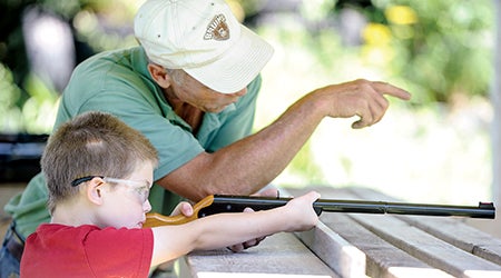 Five-year-old Anthony Schnable looks down the sight of a BB gun as Paul Jenkins helps him with aiming.